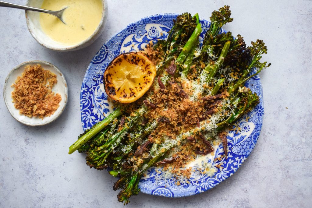 A blue plate with charred broccoli with bowls of caesar dressingf and toasted breadcrumbs on the side.