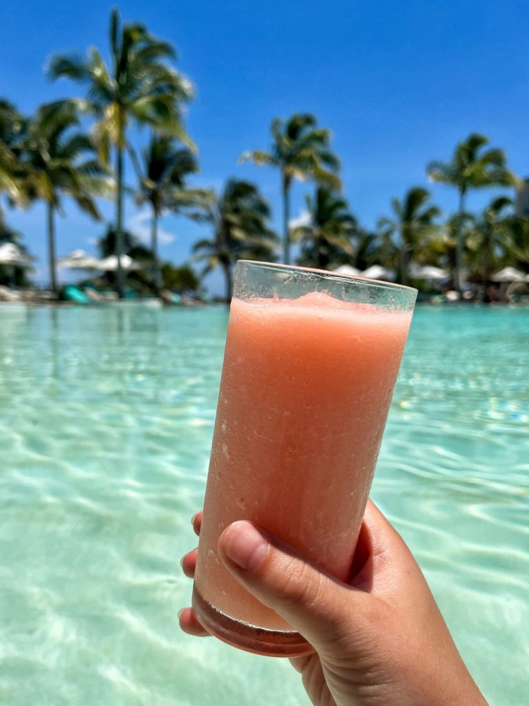 Hand holding a frozen pale pink cocktail up by a swimming pool.