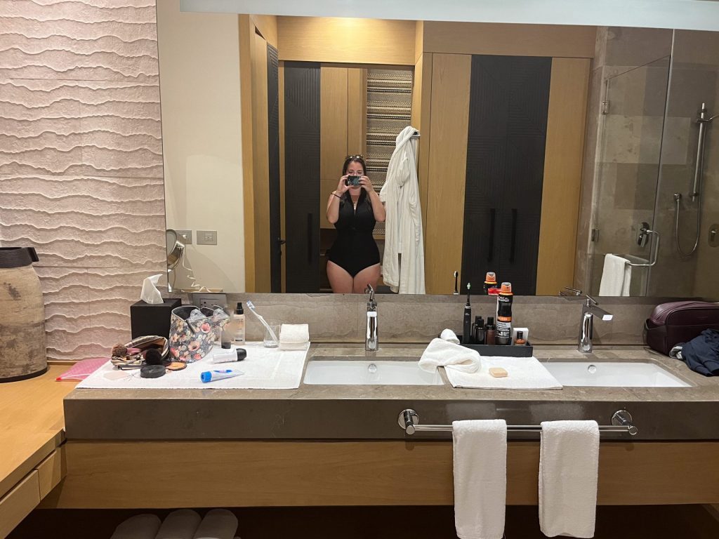 Woman in a black swimsuit taking a picture in a bathroom mirror.