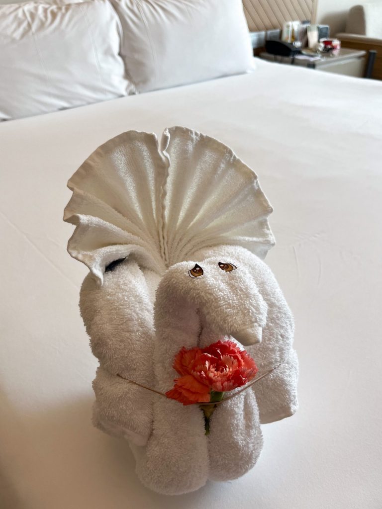 A towel swan holding a red carnation on a made bed.