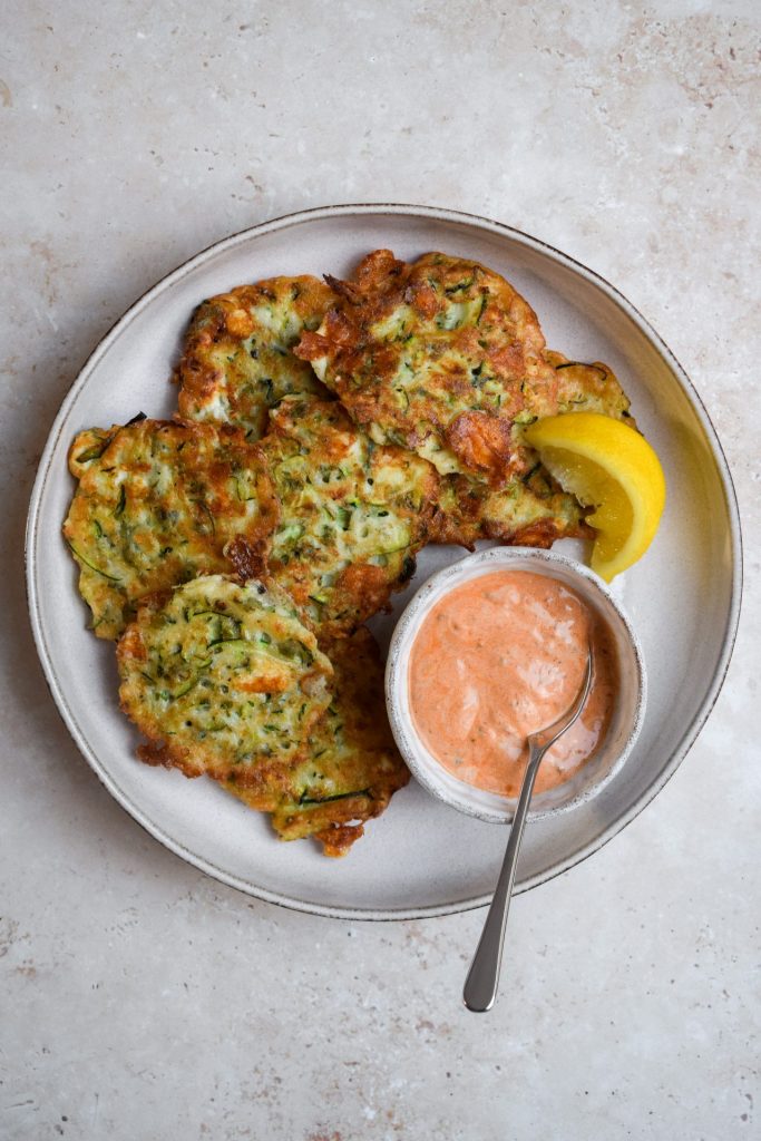 Plate of courgette fritters with a bowl of creamy orange dip and a lemon wedge.