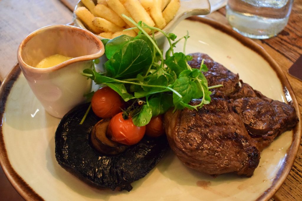 Steak, chips, mushroom, watercress and tomatoes on a plate with a jug of yellow sauce.