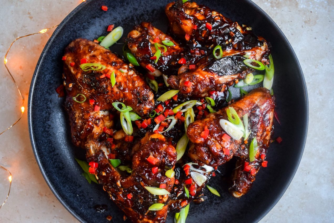 Sticky Asian Stem Ginger Chicken Wings on a black plate scattered with copped chilli and spring onions.