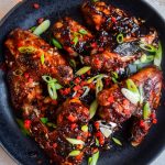 Sticky Asian Stem Ginger Chicken Wings on a black plate scattered with copped chilli and spring onions.