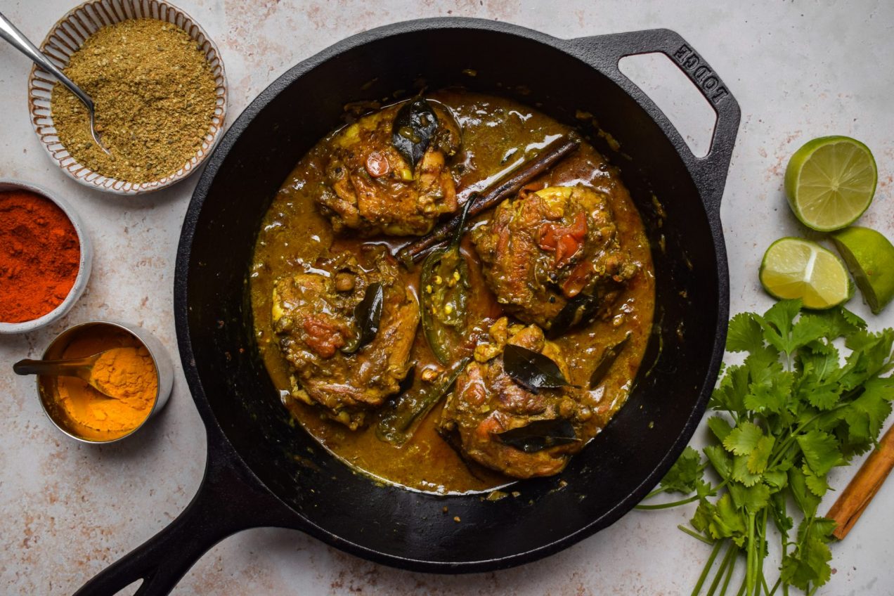 Cast iron skillet of chicken curry surrounded by spices and aromatics.
