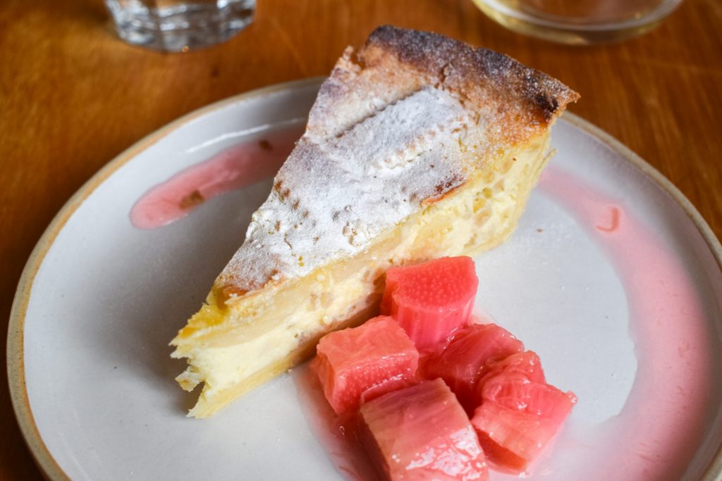 Slice of ricotta tart on a grey plate with poached rhubarb on the side.