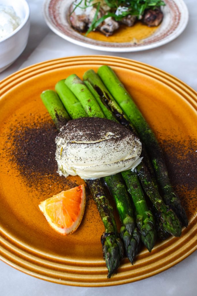Charred asparagus spears on an orange plate with a quenelle of koji butter and a small segment of blood orange.