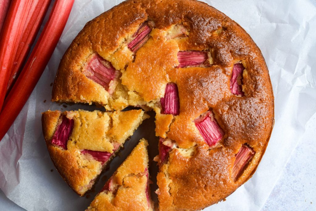 Close up of a rhubarb and white chocolate cake with stems of forced rhubarb on the side.