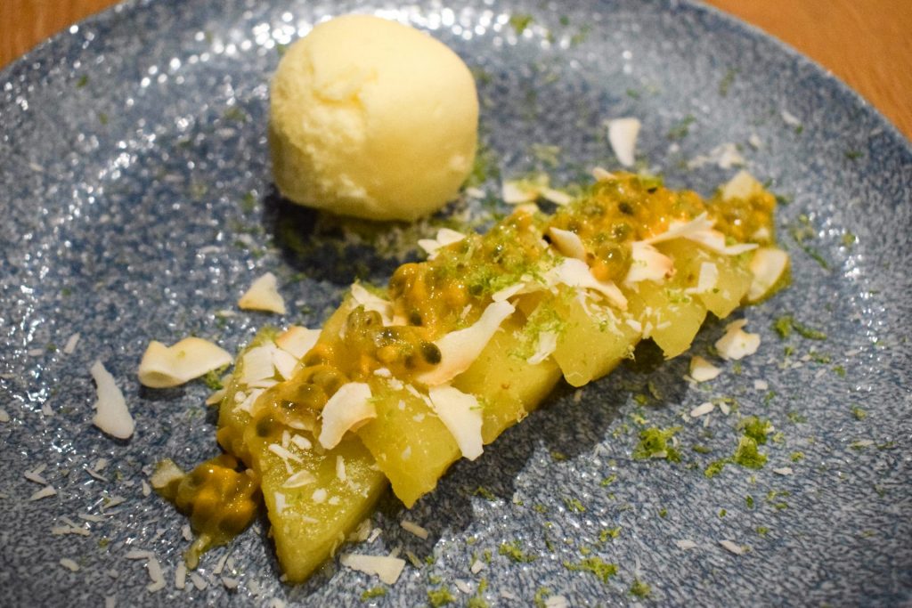 Sliced pineapple topped with passion fruit and toasted coconut with a scoop of sorbet in the background.