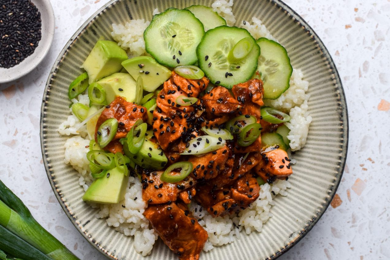 Close up of a rice bowl topped with spicy salmon, cucumber, avocado, spring onions and sesame seeds.