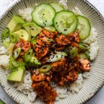 Close up of a rice bowl topped with spicy salmon, cucumber, avocado, spring onions and sesame seeds.