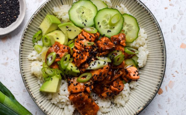 Close up of a rice bowl topped with spicy salmon, cucumber, avocado, spring onions and sesame seeds.