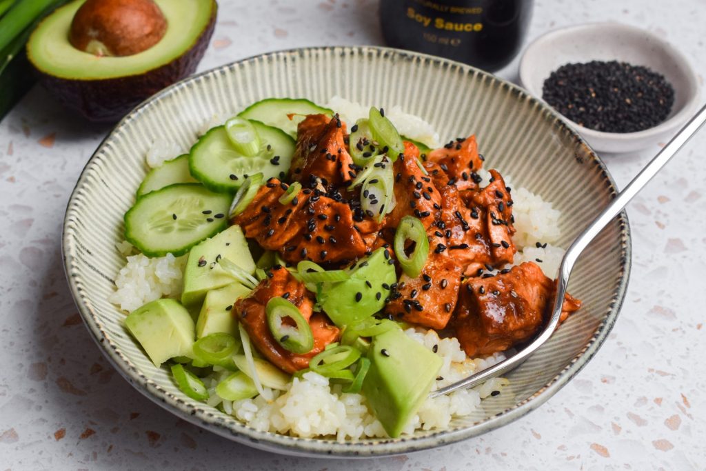 Spicy fish rice bowl topped with avocado, spring onions and sesame seeds with half an avocado and a bottle of soy sauce in the background.