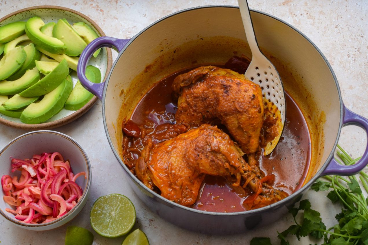 Two Yucatan Chicken legs in a purple dutch oven next to bowls of fixings.