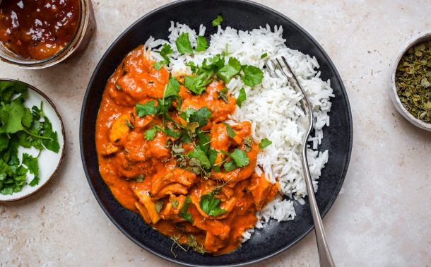 Black bowl of butter chicken and rice next to bowls of coriander, mango chutney and dried fenugreek.