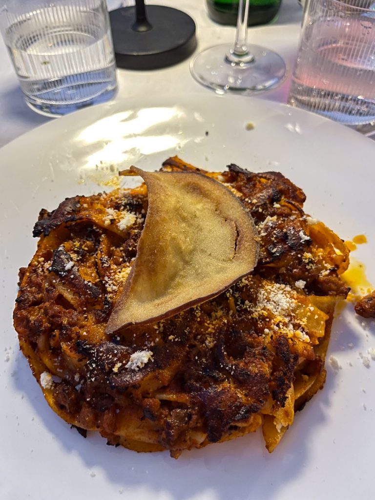 Pasta baked with ragu in a round on a white plate topped with a fried aubergine chip.