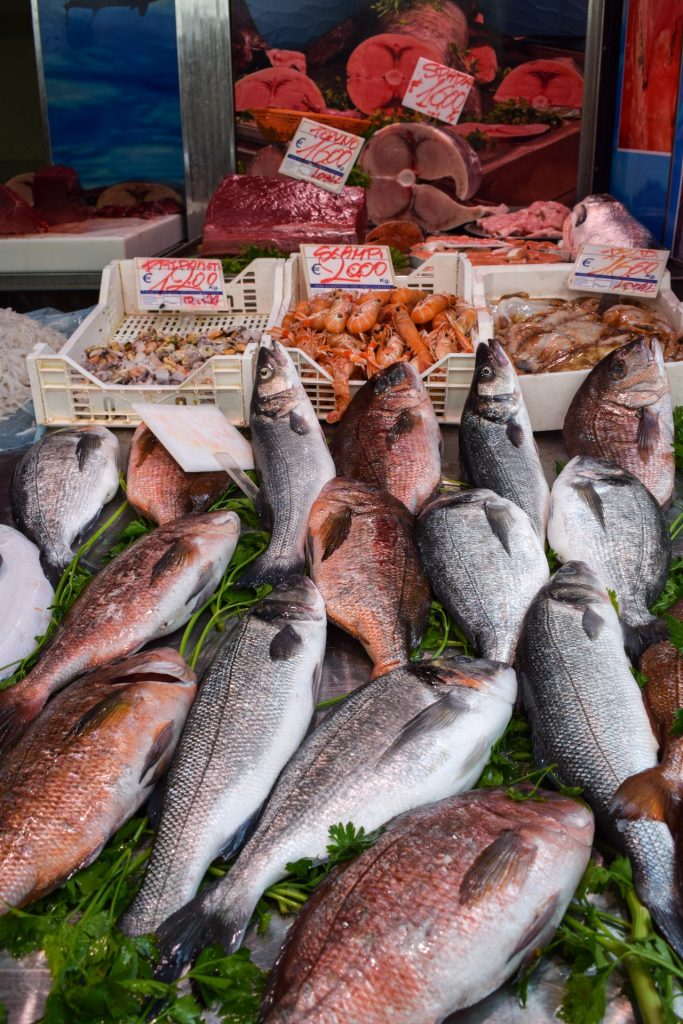 Whole fish on a market display with baskets of crustaceans and tuna steaks in the background.
