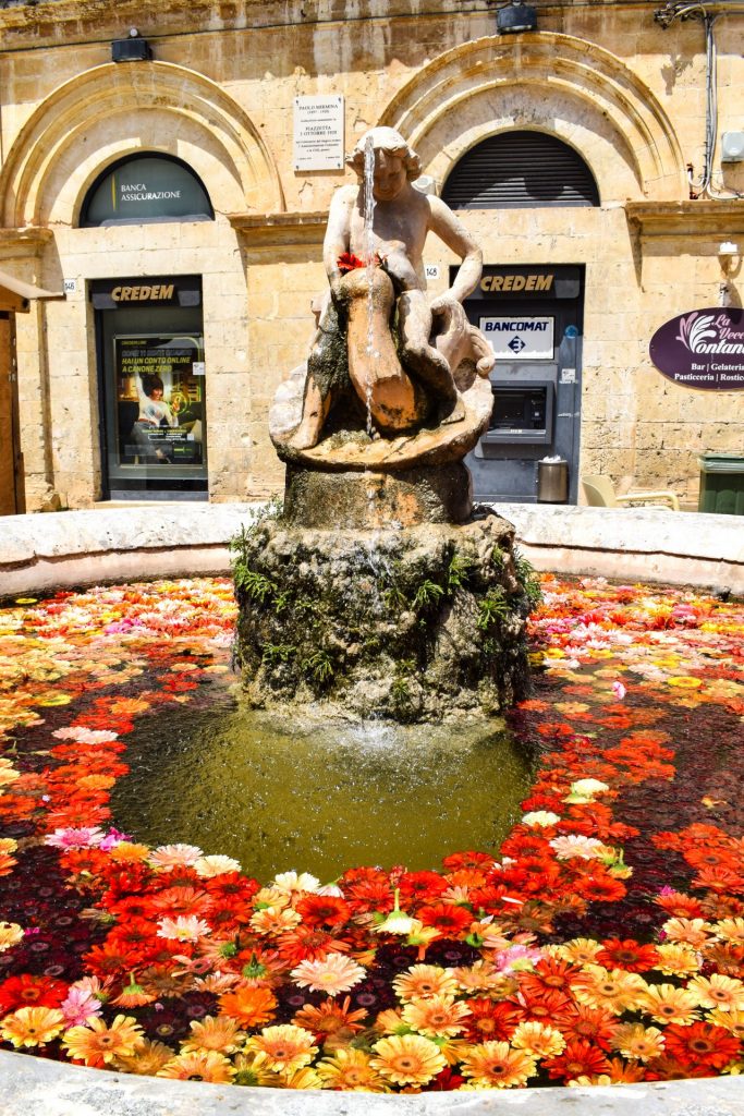 Statue of a woman in a fountain filled with floating red, orange and yellow flowers.