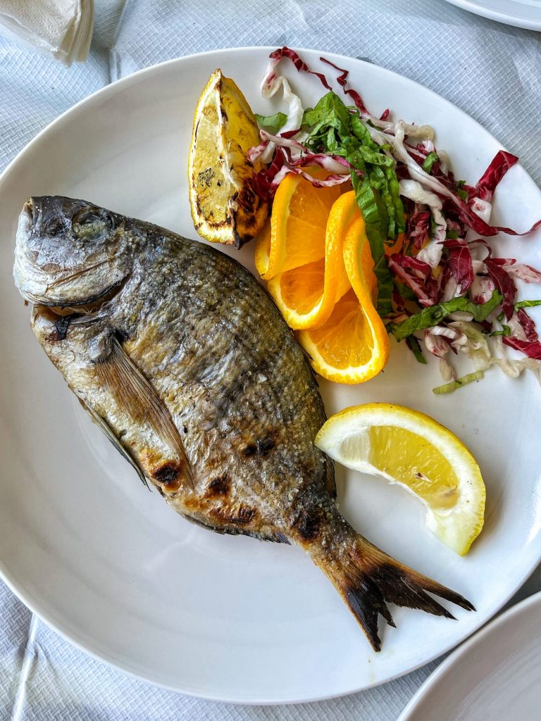 Whole grilled sea bream on a white plate with slices of orange and a small salad.