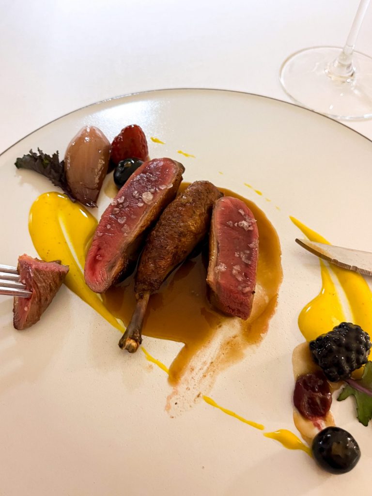 Sliced pigeon arranged on a plate with yellow and brown sauces, and berries.