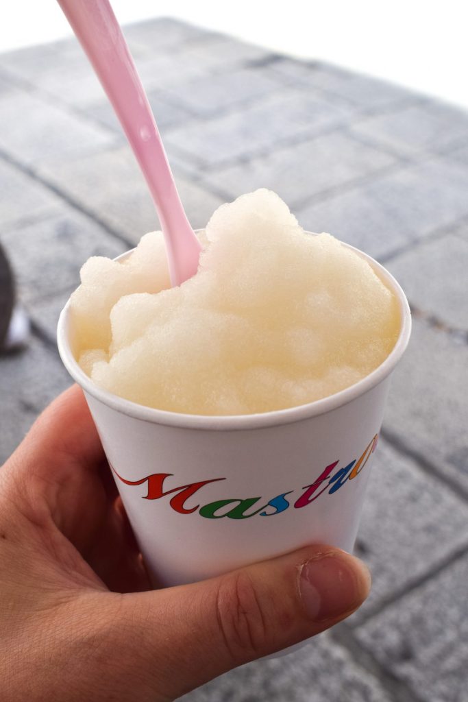 Hand holding a paper cup of lemon granita with a pink spoon.