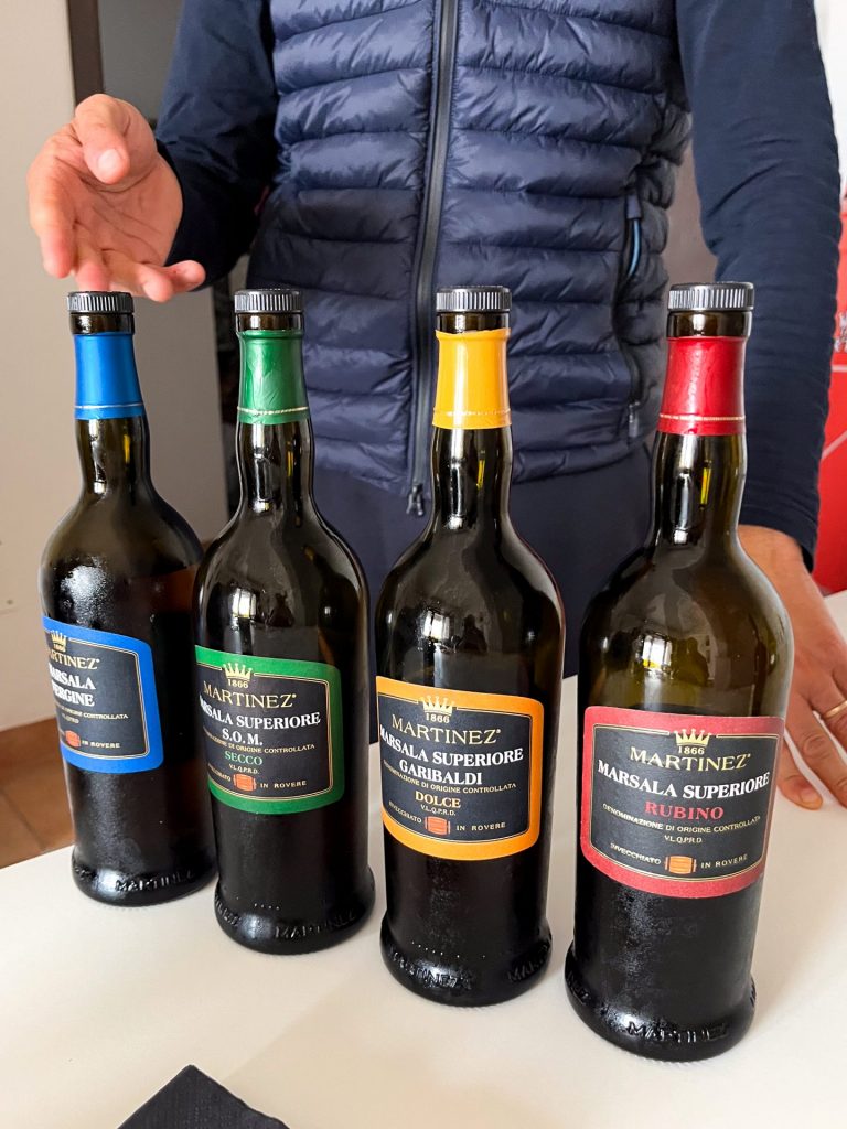 The torso of a man gesturing to four bottles of marsala on a white table.