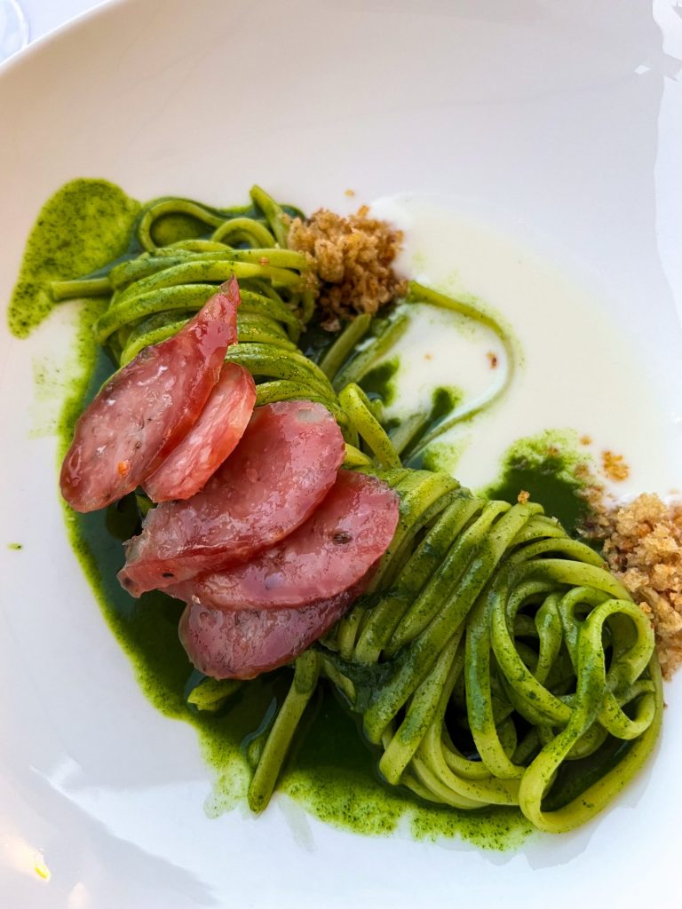 Tangle of pasta in a green sauce sitting in a puddle of cheese and topped with bright pink sliced sausage.