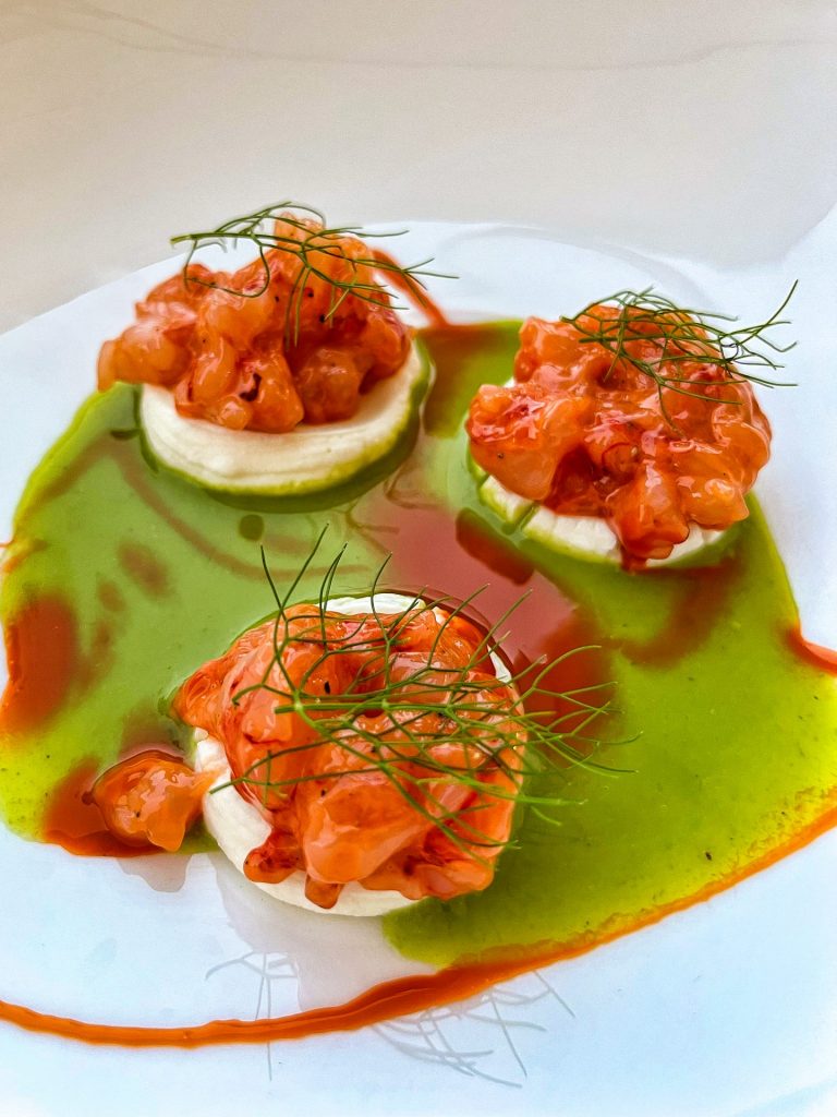 Three piles of red prawn tartare on top of piped ricotta sitting in a pool of vibrant green wild fennel oil.