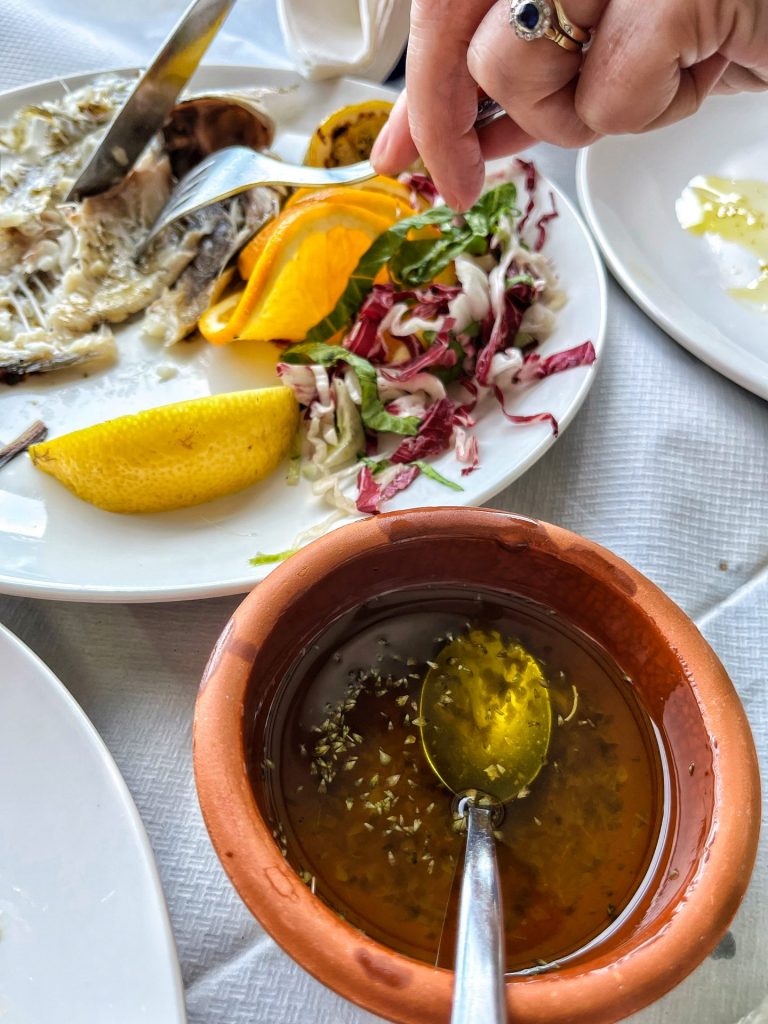 Bowl of lemon juice, olive oil and dried oregano next to a person's hands eating grilled sea bream.