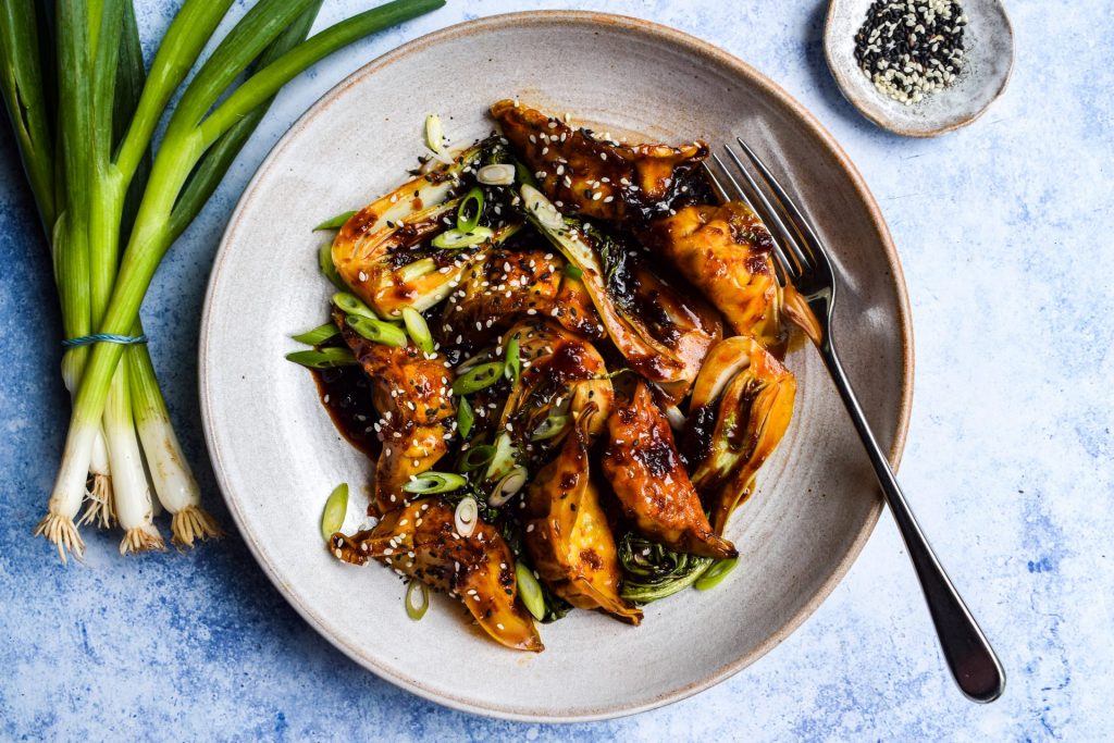 Stir fried gyoza and baby pak choi in a soy-based stir fry sauce in a stone bowl on a blue background.