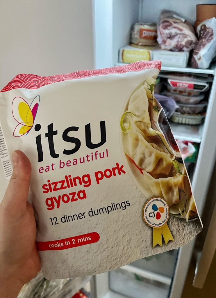 A bag of Itsu Sizzling Pork Gyoza being held up in front of an open freezer. 