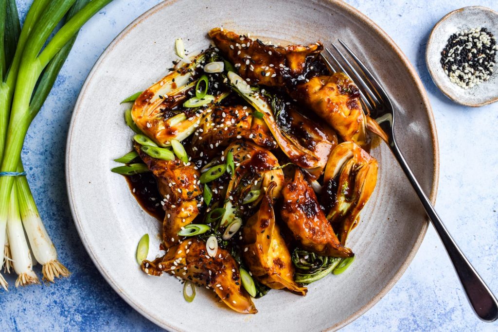 Bowl of dumplings in a dark stir fry sauce with cooked baby pak choi. Bowls of sesame seeds and a bunch of spring onions are on the side.