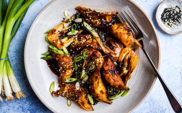 Bowl of dumplings in a dark stir fry sauce with cooked baby pak choi. Bowls of sesame seeds and a bunch of spring onions are on the side.