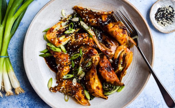 Bowl of dumplings in a dark stir fry sauce with cooked baby pak choi. Bowls of sesame seeds and a bunch of spring onions are on the side.