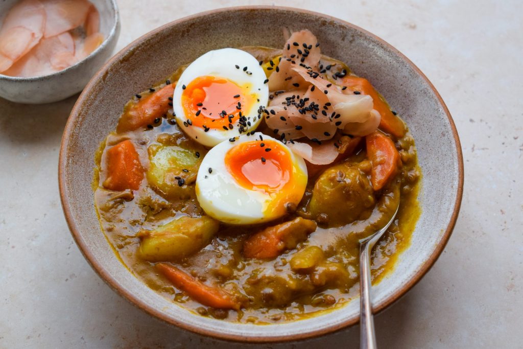 Bowl of lentil curry with potatoes, carrots and a soft boiled egg, garnished with sushi ginger and black sesame seeds.