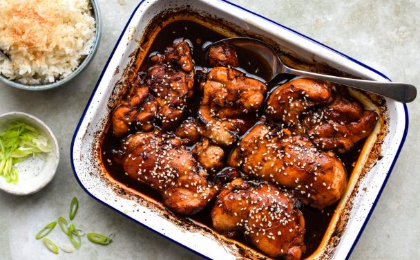 Blue and white dish of baked chicken thighs in honey and soy sprinkled with sesame seeds. A bowl of coconut rice and dish of sliced spring onions is off to one side.
