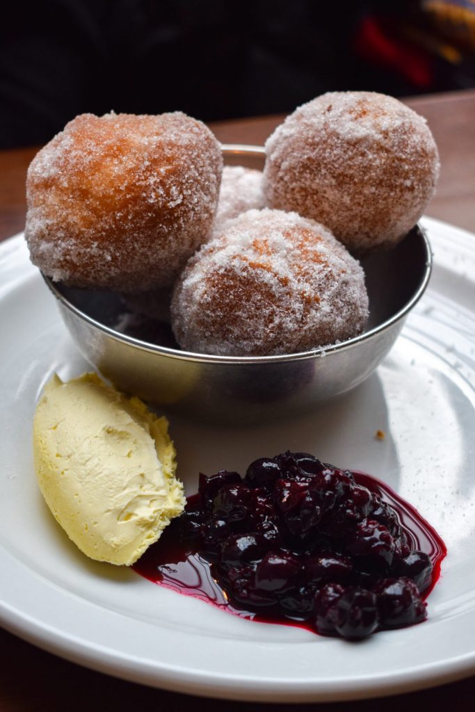 Four doughnut balls in a metal pot on a white plate, with spoonfuls of clotted cream and berry compote in the foreground.
