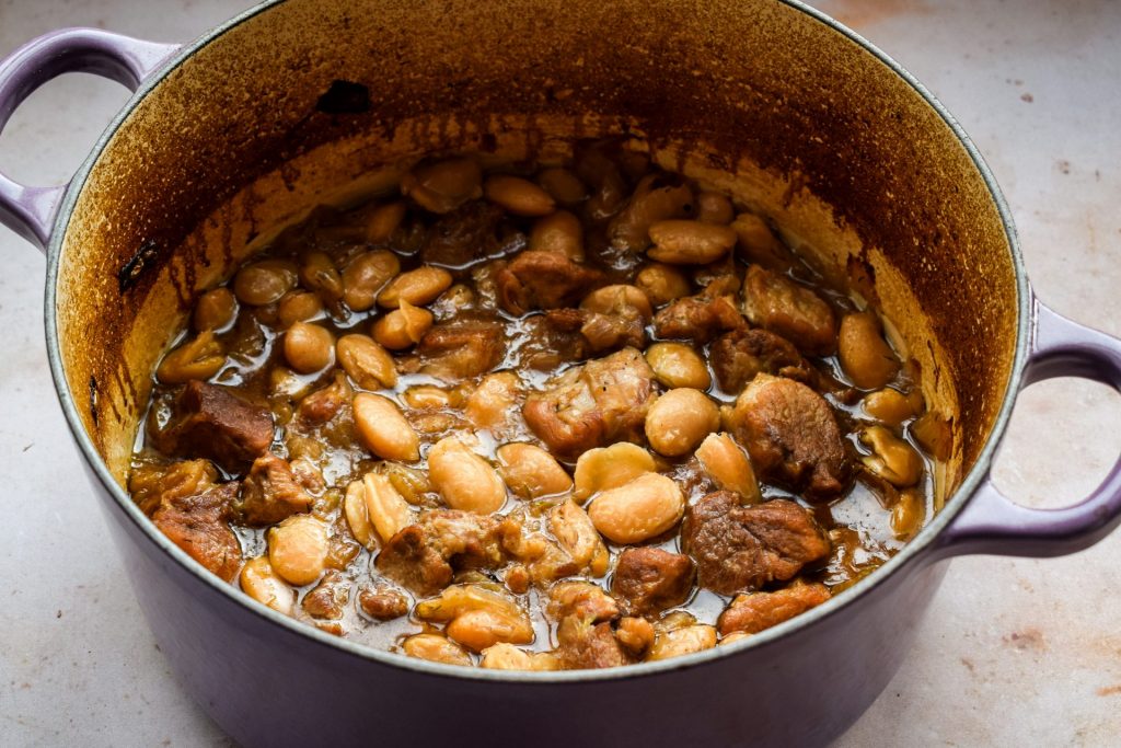 Close up of slow cooked pork and beans in sherry pictured in a purple Le Creuset casserole dish.