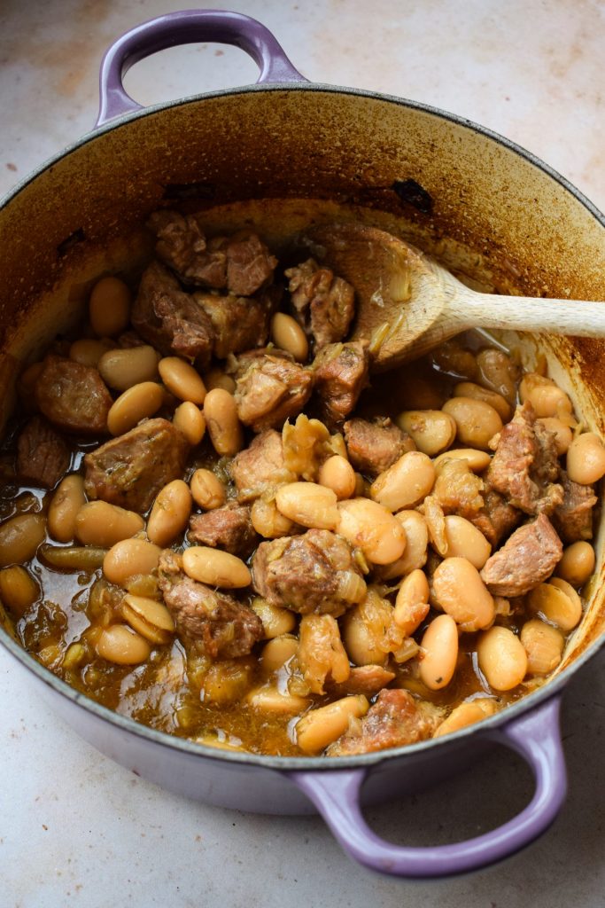 Slow cooked pork with butter beans being stirred with a wooden spoon inside a purple casserole dish.