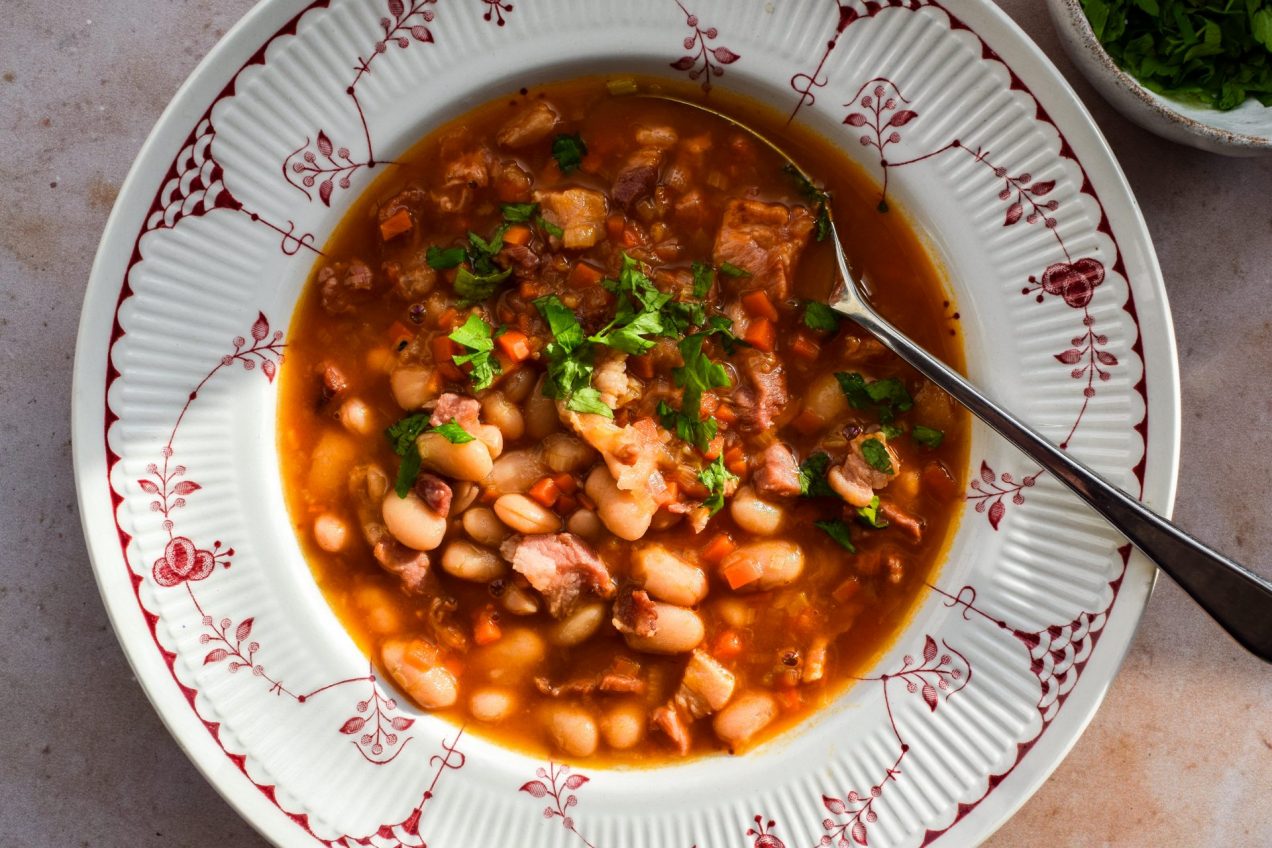 Close up of a red and white patterned bowl with bean and bacon soup topped with chopped parsley.