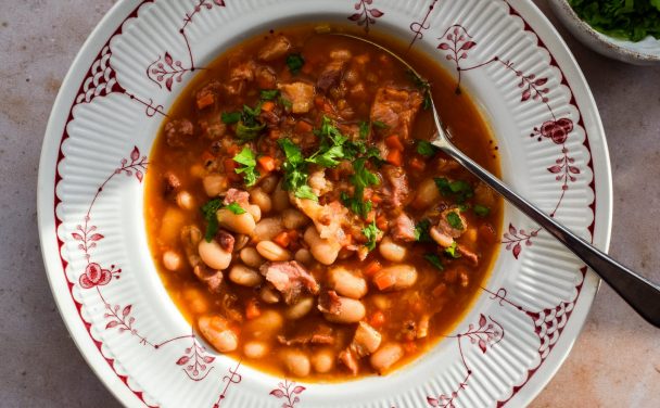 Close up of a red and white patterned bowl with bean and bacon soup topped with chopped parsley.