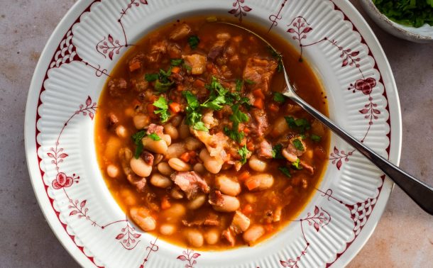 Close up of a red and white patterned bowl with bean and bacon soup topped with chopped parsley.