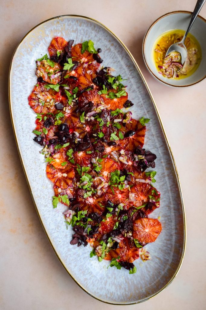 Large oval serving platter of blood orange salad with parsley, chopped black olives, and a bowl of dressing off to one side.