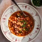 Red and white bowl of bacon and white bean soup with a bowl of flat leaf parsley for scattering off to one side.