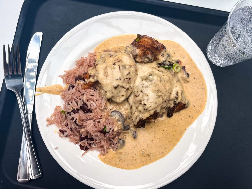Plate of jerk chicken thighs with beans, rice and a cream sauce on a white plate on a cafeteria tray.
