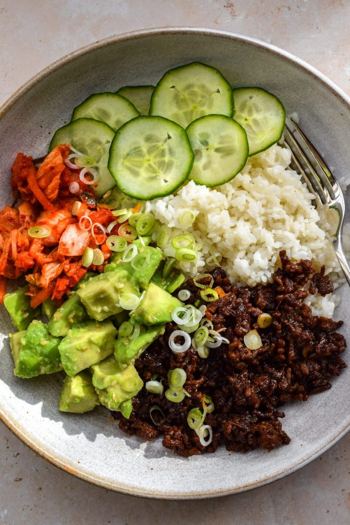 Korean beef bowl close up with seasoned mince, avocado salad, kimchi, cucumber and spring onions.