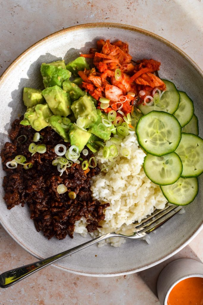 Close up of a sticky rice bowl with sliced cucumber, korean beef mince, kimchi and avocado tossed with sesame seeds.