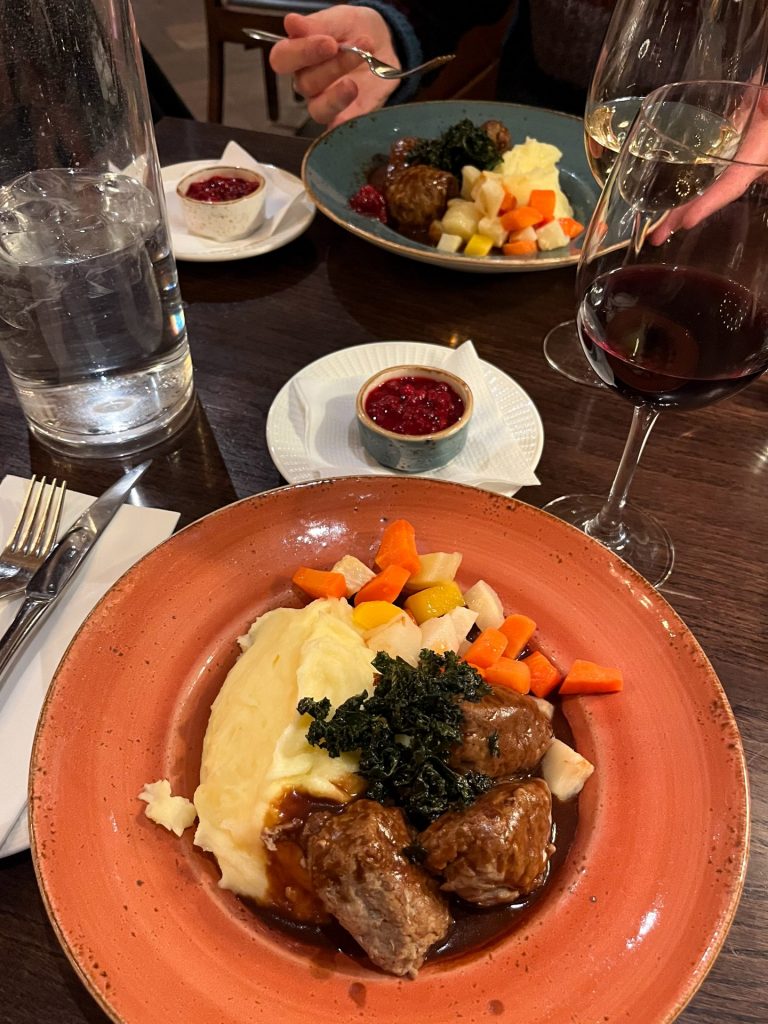 Orange plate of Norwegian meatballs, mashed potatoes and vegetables with another serving and pots of lingonberry jam in the background.