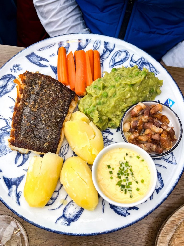 Place of pan fried fish, boiled potatoes, carrots and mushy peas with small pots of white wine sauce and crispy bacon lardons.