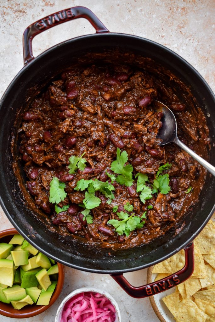 Overhead view of a pot of beef short rib chili with coriander leaves scattered on top. Avocado, pickled onions and tortilla chips are off to one side.